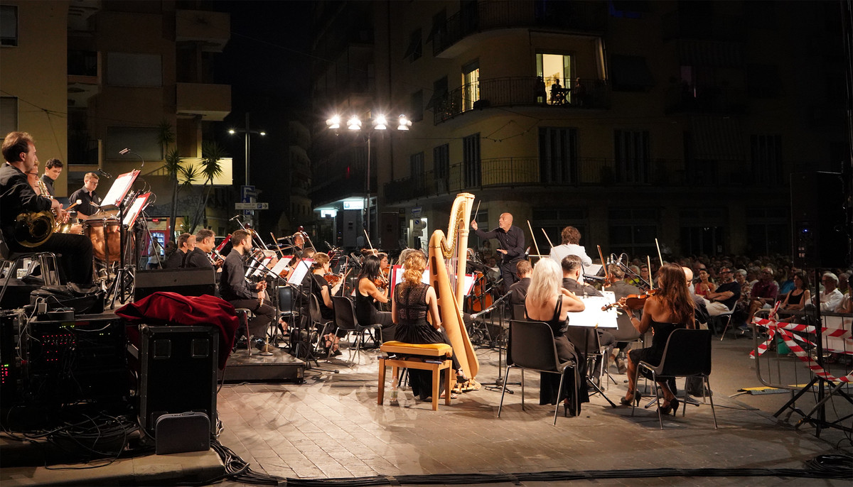 Piazza di un borgo storico, pubblico seduto, orchestra con strumenti a fiato e archi durante esibizione serale estiva