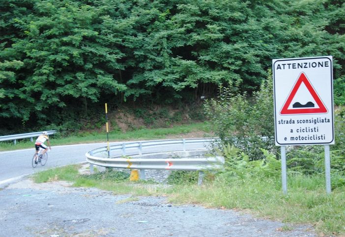 Il cartello stradale con, sullo sfondo, un ciclista che affronta in discesa la prima curva della "Colletta" di Barge